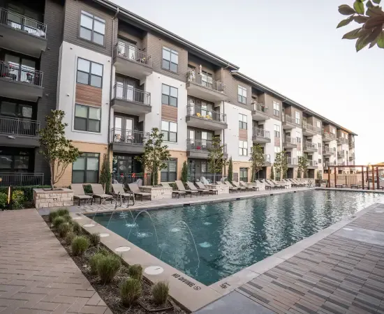 A modern four-story HOA-managed apartment community with a long resort-style swimming pool, lounge chairs, paver walkways, manicured plantings, and architectural facades in late-day light. The kind of commercial maintenance property a landscape account manager walks during a SiteDNA pre-inspection before submitting a competitive bid to the HOA board.